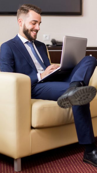 Full length portrait of handsome young businessman using laptop in hotel room and smiling while relaxing casually in armchair