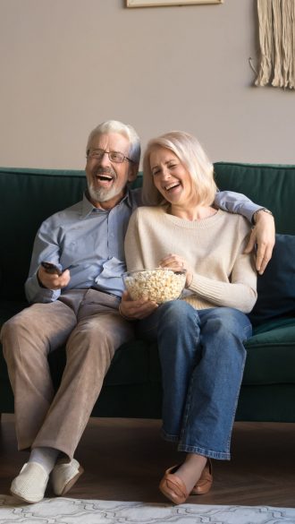 Happy aged couple, wife and husband watching tv, comedy movie and eating popcorn snack, laughing, sitting on cozy couch at home, mature family, man and woman enjoying free time, weekend together