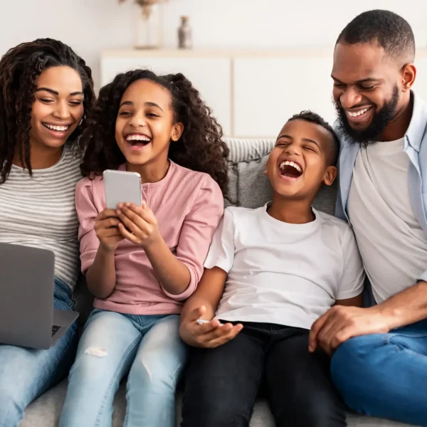 Entertainment And Leisure. Portrait of happy African American parents and their little children using laptop and smartphones, spending time together sitting on the couch in living room and laughing